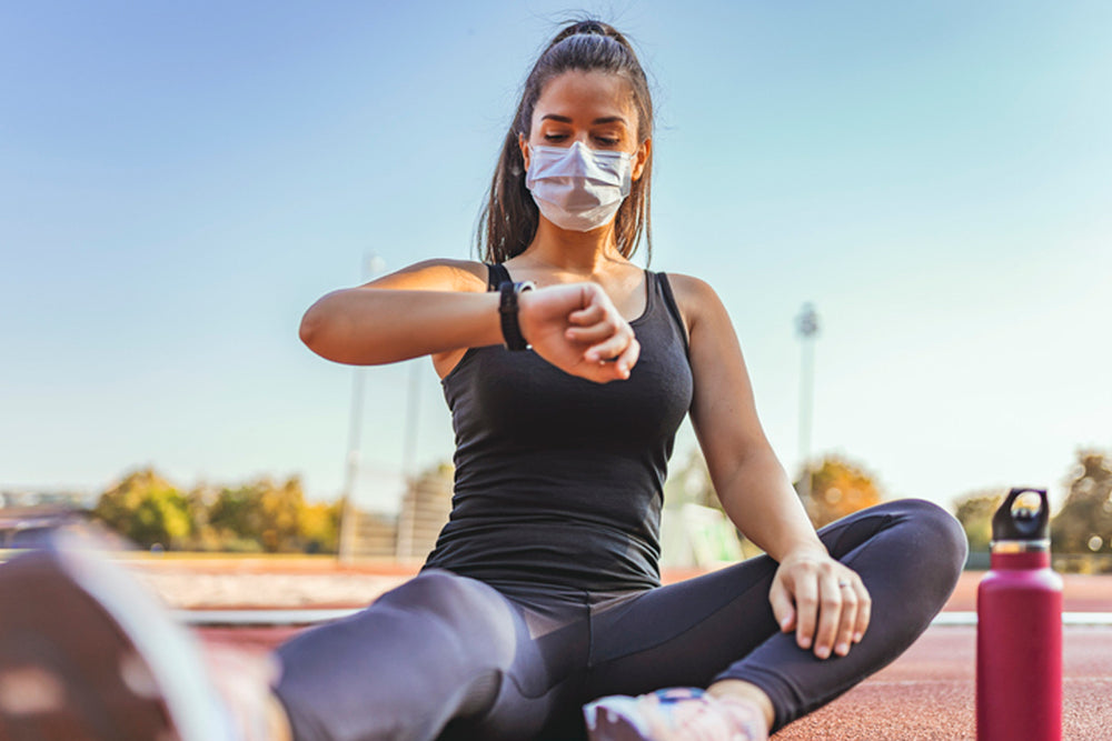 Woman in exercise clothes and a face mask checking her watch.