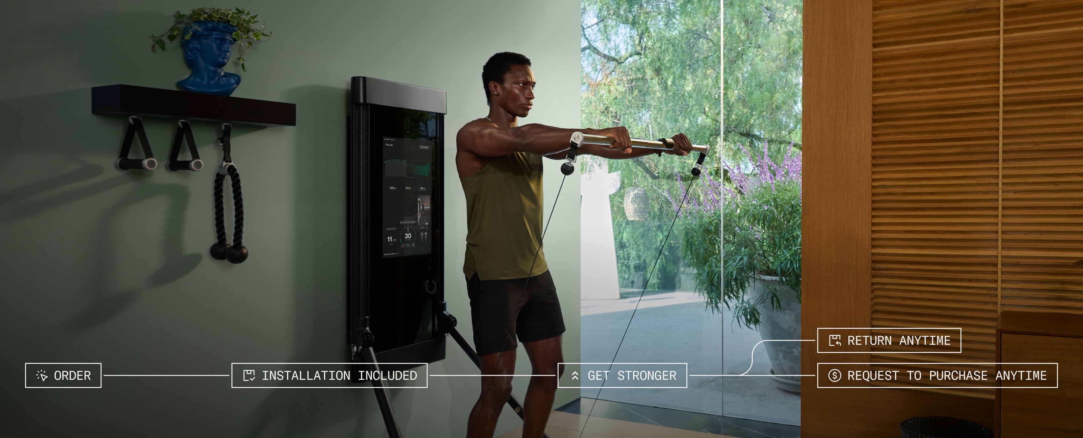 Man exercising using a wall-mounted exercise machine, Tonal, in a home setting with greenery outside.