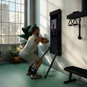 Person performing a squat with the Tonal Bar and Bar Control Module with a Tonal fitness device displaying an app interface.