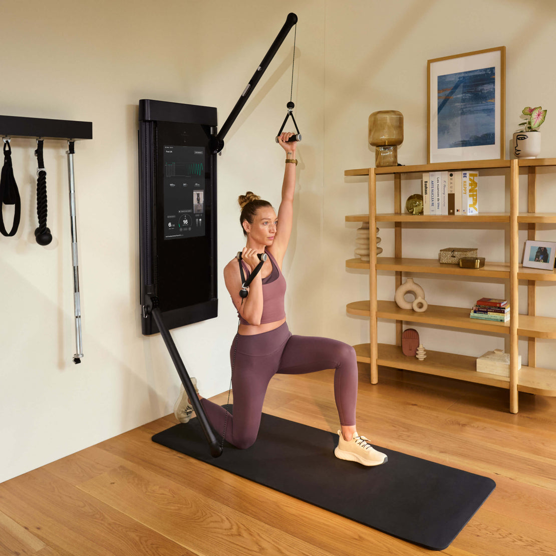 Woman performing a lunge and overhead press with a Tonal fitness device displaying an app interface.