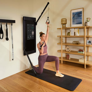 Woman performing a lunge and overhead press with a Tonal fitness device displaying an app interface.