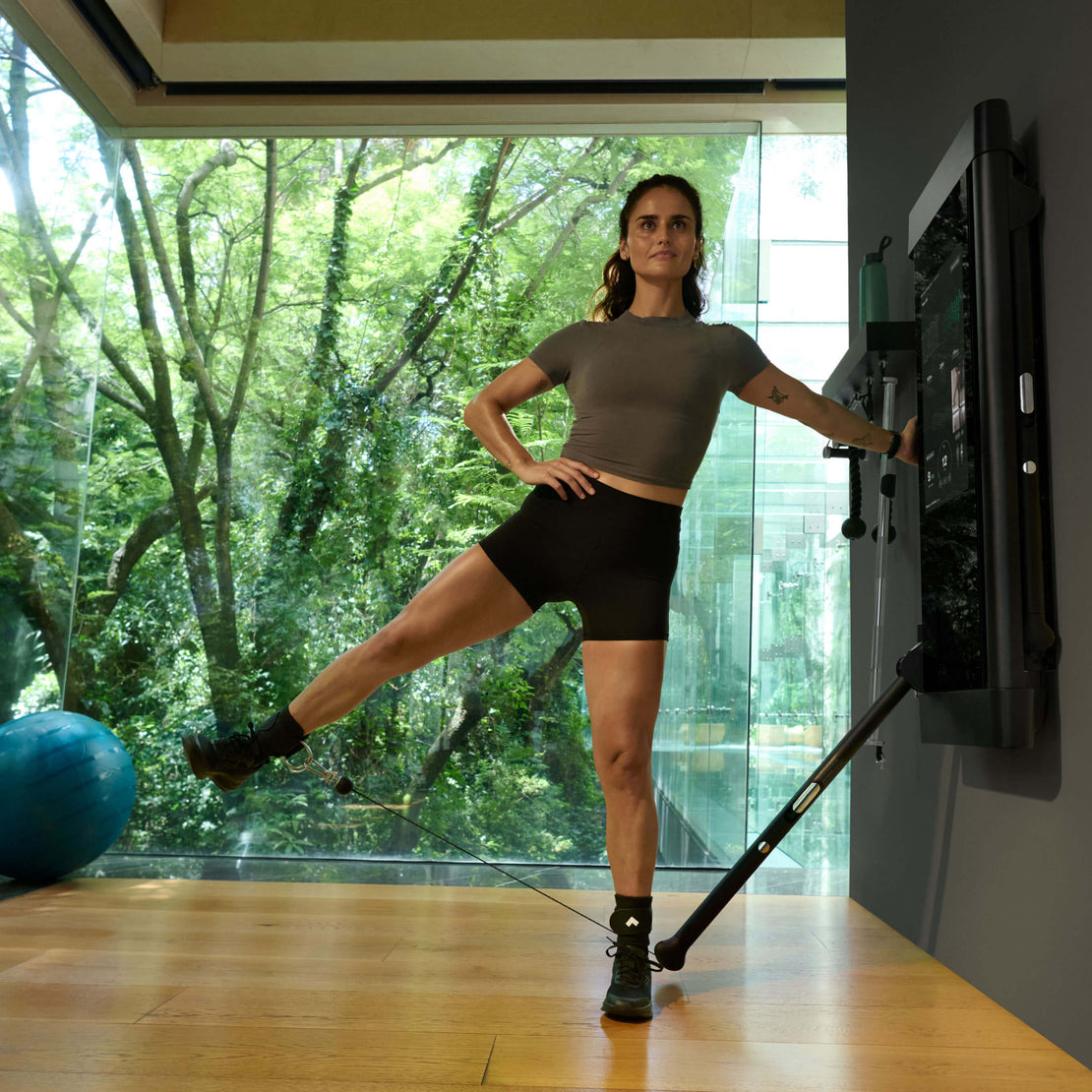 Woman exercising with Tonal Ankle Straps on Tonal 2 in a room with large windows showing greenery outside.