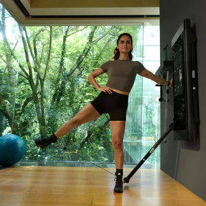 Woman exercising with Tonal Ankle Straps on Tonal 2 in a room with large windows showing greenery outside.