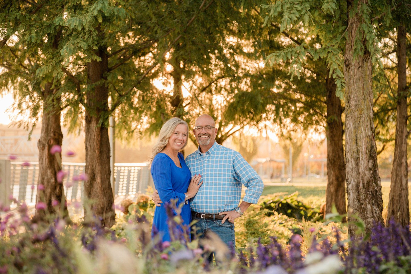 a woman and man stand holding each other and smiling surrounded by trees and flowers